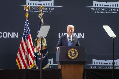 President Joe Biden speaking at The Dodd Center for Human Rights at the University of Connecticut.  President Biden is standing at a podium featuring the U.S. Presidential seal with American flags in the background