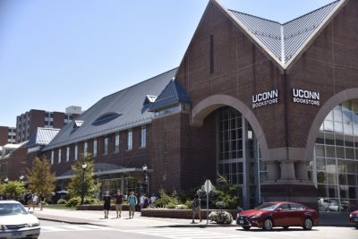Exterior view of the UConn Bookstore building which is a large brick building at the corner of an intersection. Students walking on a sidewalk and cars driving through the intersection are in the foreground