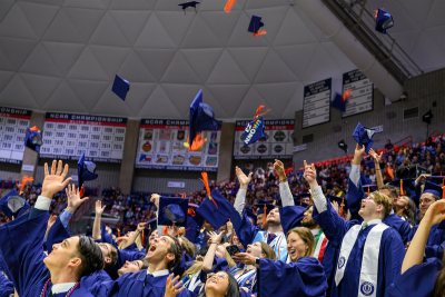 Crowd of graduates in blue and white regalia at a commencement ceremony in Gampel Pavilion tossing their caps in celebration