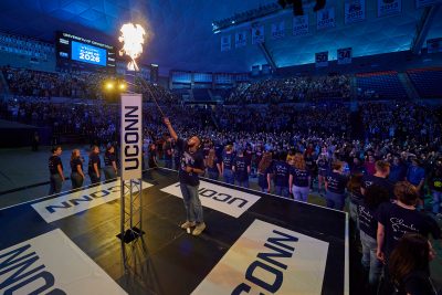 Individual lighting a large ceremonial torch on a stage surrounded by a crowd of students in Gampel Pavilion at the Class of 2026 Convocation event