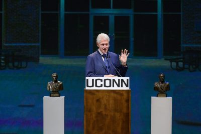 Former President Bill Clinton speaking at a UConn podium in front of the Dodd Center flanked by Dodd Prize busts on pedestals