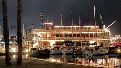 A riverboat in port lit up at night in San Diego, California.  A variety of smaller sailboats are alongside the riverboat with a small beach in the foreground