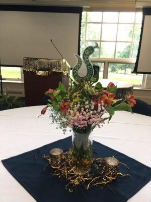 Floral centerpiece on a banquet table featuring a paper snake decoration and gold streamers and votives around the vase. There is a podium with gold tassels around it in the background