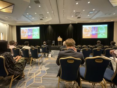 Professionals seating in the audience listening to a speaker at a podium.  The speaker is referencing images displayed on monitors on either side of the podium